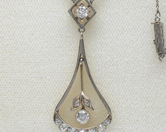 May include: A silver-toned pendant necklace with a diamond-shaped top and a teardrop-shaped pendant. The pendant features a large central diamond, smaller diamonds, and a floral design. The necklace is set against a light background.