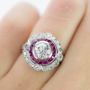 Puede incluir: Primer plano de un anillo con una gran piedra central de diamante transparente y redondo. El anillo tiene un halo de diamantes más pequeños y un anillo de piedras rojas. El anillo está en un dedo.