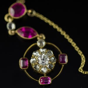 May include: A gold necklace with a central pendant featuring a large, clear gemstone surrounded by smaller, clear gemstones. The necklace also includes pink gemstones and a delicate gold chain.