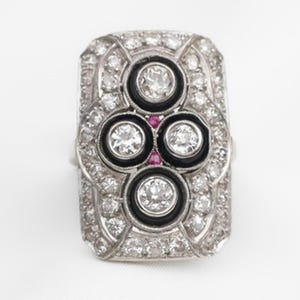 Può includere: Un anello rettangolare in stile Art Déco con montatura in metallo color argento. L'anello presenta un design centrale di quattro pietre rotonde e chiare incastonate in smalto nero, con piccole pietre rosse tra di esse. L'anello è incrostato con numerose pietre chiare più piccole.