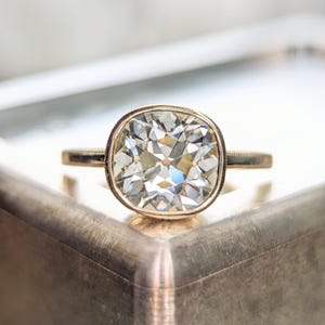 May include: A gold ring featuring a large, square-cut diamond. The diamond is set in a bezel setting and has a brilliant, multifaceted appearance. The ring's band is thin and simple, with delicate detailing near the diamond.