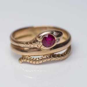 Puede incluir: Anillo de serpiente dorado con un ojo de rubí y dos pequeños diamantes. El anillo se enrolla alrededor del dedo. La cabeza de la serpiente está en la parte superior y la cola en la inferior.