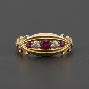 May include: A gold ring with a decorative design, featuring three round, vibrant red gemstones and two clear, round gemstones. The ring has an ornate, flowing design with a polished finish, set against a dark background.