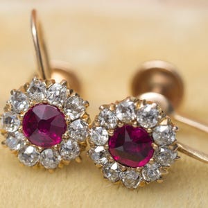 May include: A pair of gold earrings with a central, deep red gemstone encircled by smaller, clear stones. The earrings have a screw-back design. The jewellery is set against a soft, yellow background.