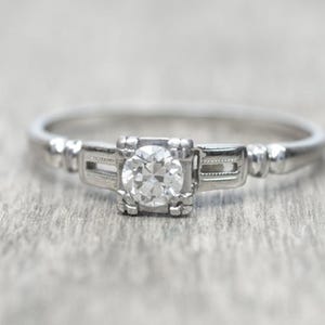 May include: A close-up of a silver ring with a round, clear diamond center stone. The ring has a detailed setting with rectangular accents on either side of the diamond. The band is thin and polished.