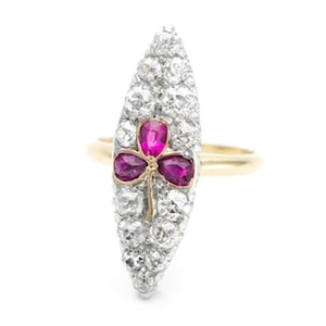May include: A gold ring with a marquise-shaped face, encrusted with small diamonds. The center features a three-leaf clover design with three vibrant red gemstones. The ring is set against a white background.