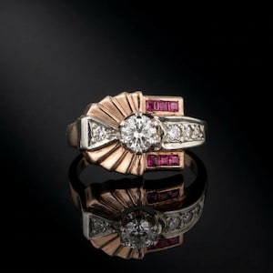 May include: A two-tone gold and silver ring with a large, round, clear diamond centre stone. The ring features smaller diamonds and red stones, with a fan-like design around the centre stone. The ring is set against a black background.