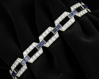 Op de afbeelding: Een zilveren armband met een geometrisch ontwerp. De armband heeft rechthoekige blauwe edelstenen en heldere, ronde edelstenen. De armband wordt op een zwarte achtergrond gepresenteerd.