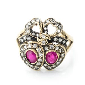 May include: A gold-toned ring featuring a double heart design with a bow detail. Each heart contains a vibrant pink gemstone, surrounded by small, sparkling white stones. The ring has a vintage aesthetic.