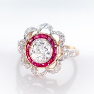 May include: A vintage-style ring with a large, clear center stone surrounded by a halo of small, sparkling diamonds. The center stone is encircled by a row of red gemstones, set in a gold and silver band.