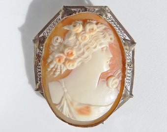 May include: A cameo brooch featuring a carved portrait of a woman with a floral crown, set in a silver filigree frame.