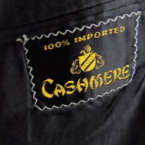 Op de afbeelding: Een zwarte stoffen kledingvoering met een rechthoekig zwart label dat is vastgezet met witte zigzagstiksels. Het label toont de geel geborduurde tekst 100% IMPORTED CASHMERE en een klein geel wapenschild met een pluim erboven.