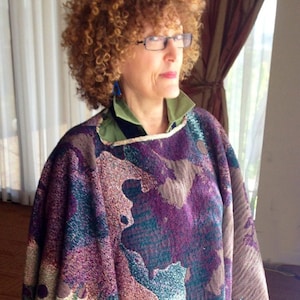 May include: A woman wearing a colorful woven poncho with a fringe border. The poncho has a purple, blue, green, and brown pattern. The woman is wearing blue jeans and black shoes. The poncho has the text "Diane Bush" woven into the fabric.