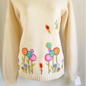 May include: A cream-colored knitted sweater with colorful embroidered flowers. The flowers are in shades of pink, blue, green, orange, and yellow.