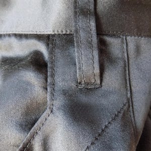 May include: Close-up of black fabric with visible stitching and a belt loop. The fabric has a subtle sheen, suggesting a satin or silk-like material. The stitching is precise, highlighting the garment's construction.