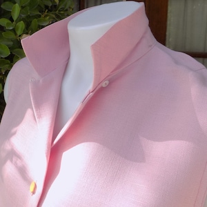 May include: A sleeveless pink button-up shirt with a collar. The shirt is made of a textured fabric and has a single button closure at the neck.
