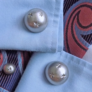 May include: Two silver cufflinks with a star design on a light blue fabric background. The cufflinks are round and have a shiny finish.