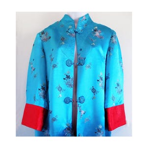 Pode incluir: Uma jaqueta de seda azul turquesa com gola mandarim e acabamento vermelho nas mangas. A jaqueta tem um padrão floral e é fechada com botões decorativos.