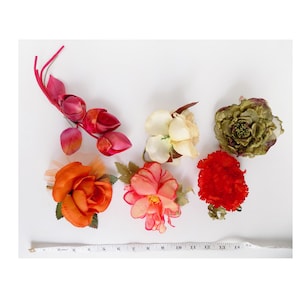 May include: Five artificial flower embellishments in various colors and styles.  A cluster of pink flower buds, a peach-colored flower, a red carnation, a cream-colored flower, and a green and brown flower.