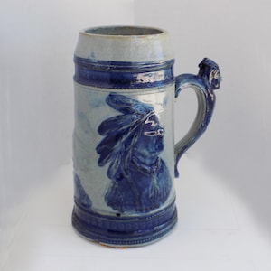 May include: A large, blue and grey ceramic beer stein with a handle. The stein features a relief of a Native American chief in profile. The handle is adorned with a sculpted face. The stein has a lid and is decorated with bands of blue.