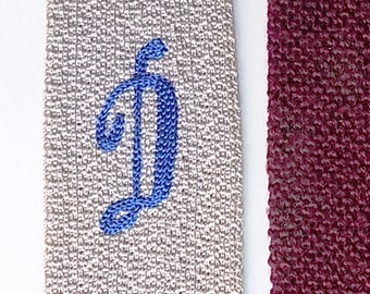 May include: A close-up of a white fabric with a blue embroidered letter "D" on the left side. The fabric is next to a dark red fabric.