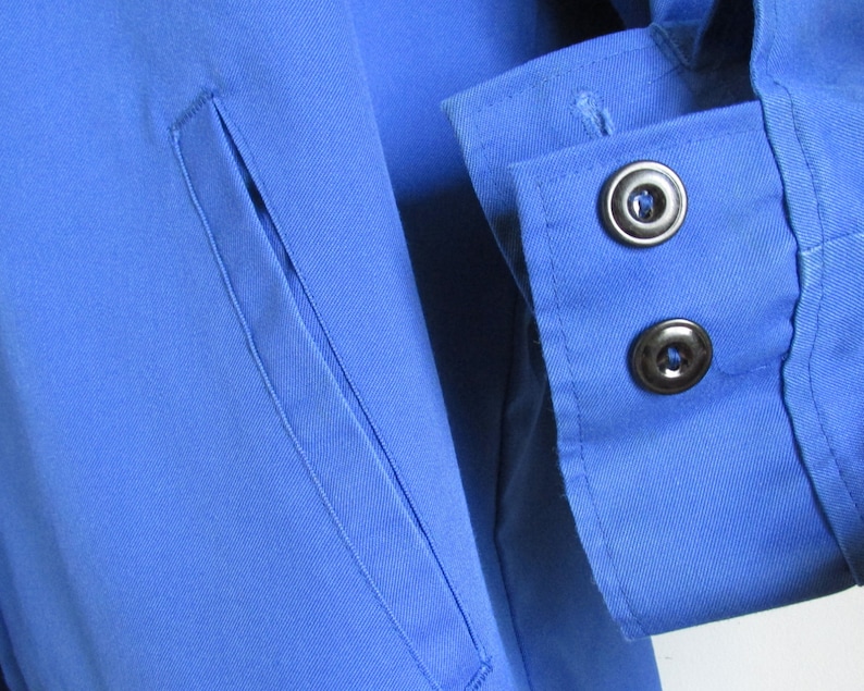 Puede incluir: Primer plano de una camisa azul con un bolsillo y dos botones negros en el pu&ntilde;o.
