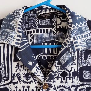 Pu&ograve; includere: Una camicia abbottonata con colletto a fantasia bianca e nera. La camicia presenta un design geometrico con figure bianche e nere su sfondo nero. L'etichetta sulla camicia recita "Made in Hawaii".