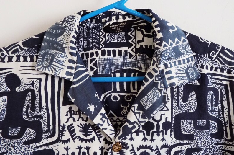 Pu&ograve; includere: Una camicia abbottonata con colletto a fantasia bianca e nera. La camicia presenta un design geometrico con figure bianche e nere su sfondo nero. L'etichetta sulla camicia recita "Made in Hawaii".