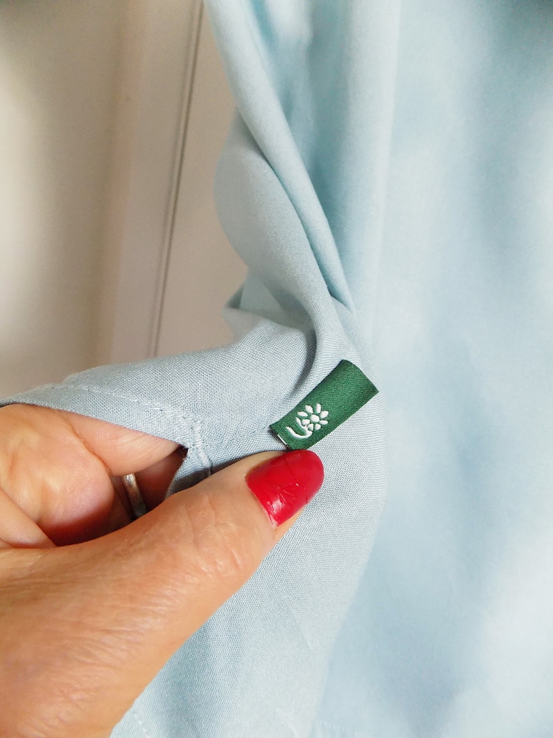 Puede incluir: Una camisa azul claro con una etiqueta verde que tiene una flor blanca.