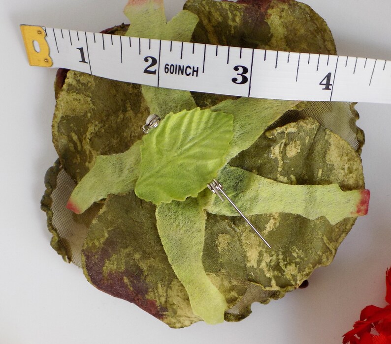 May include: A green and brown fabric rose with a green leaf and a silver pin. The rose is about 2 inches in diameter.