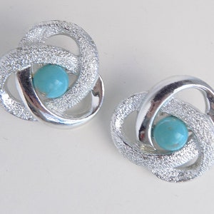 Może przedstawiać: Para srebrnych kolczyków z turkusowym kamieniem w środku każdego kolczyka. Kolczyki mają kształt węzła.