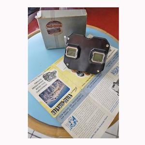 May include: A black plastic View-Master viewer with two red and two clear lenses. The viewer is sitting on a blue and white patterned surface with a folded pamphlet that reads "View-Master are precision built for years of perfect operation".