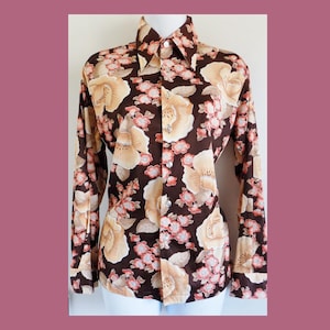 May include: A long-sleeved shirt with a brown base, featuring a large floral pattern in beige, peach, and pink. The shirt has a pointed collar and buttoned cuffs. The floral design is a repeating pattern across the fabric.
