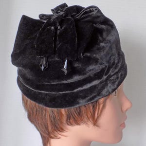 May include: A black velvet hat with a large bow on top. The hat is decorated with two black crystal-like embellishments.