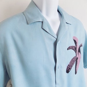 Puede incluir: Una camisa de manga corta azul claro con botones y un estampado gr&aacute;fico de pl&aacute;tano rosa y negro.