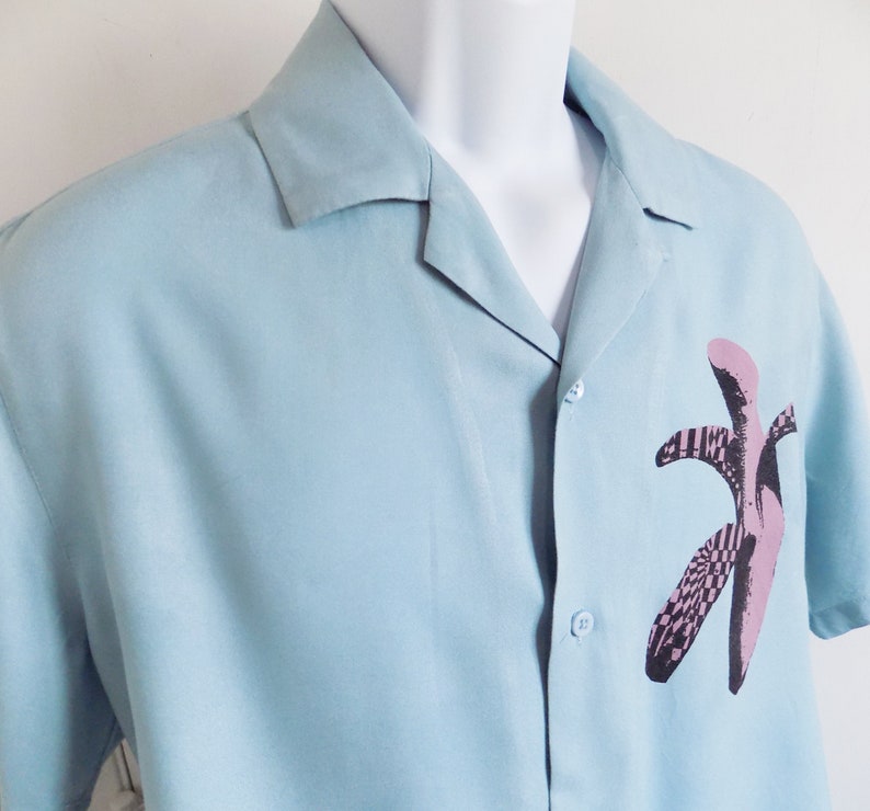Puede incluir: Una camisa de manga corta azul claro con botones y un estampado gr&aacute;fico de pl&aacute;tano rosa y negro.