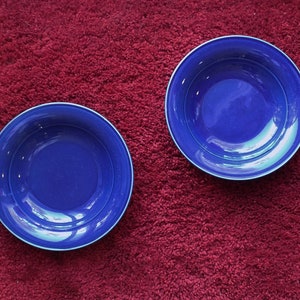 May include: Two blue ceramic bowls on a red carpet. The bowls are empty and have a glossy finish.