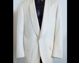 Può includere: Una giacca da smoking color bianco sporco con collo a scialle, chiusura a un bottone e due tasche con patta in vita. Il capo è esposto su un manichino e presenta maniche lunghe e una silhouette strutturata e sartoriale.
