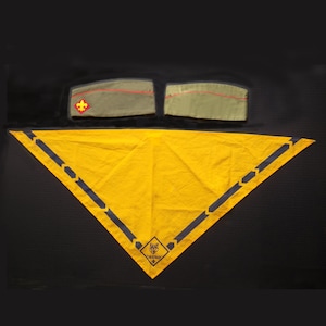 Puede incluir: Un pañuelo triangular amarillo con bordes azul oscuro y un logotipo, junto con dos gorras de explorador verde oliva con ribetes y parches rojos. Los artículos se muestran sobre una superficie negra.