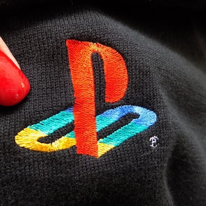 Puede incluir: Un parche de tela negro con un logotipo de PlayStation bordado. El logotipo tiene la forma de una "P" estilizada con un círculo alrededor. La "P" es roja, el círculo es azul, verde y amarillo.