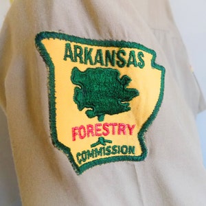 May include: A yellow and green embroidered patch with the text "Arkansas Forestry Commission". The patch is shaped like the state of Arkansas with a green tree inside.
