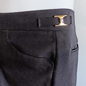 May include: Close-up of dark charcoal-colored pants with a gold-colored buckle. The pants have a tailored fit, featuring a front pocket and a small back pocket. The gold buckle adds a touch of elegance to the design.