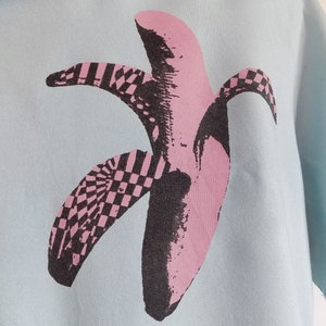 Puede incluir: Una camisa azul claro con un estampado gr&aacute;fico de pl&aacute;tano rosa y negro. El pl&aacute;tano est&aacute; estilizado con un patr&oacute;n a cuadros.