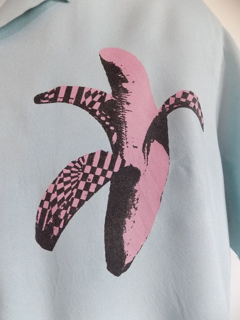 Puede incluir: Una camisa azul claro con un estampado gr&aacute;fico de pl&aacute;tano rosa y negro. El pl&aacute;tano est&aacute; estilizado con un patr&oacute;n a cuadros.
