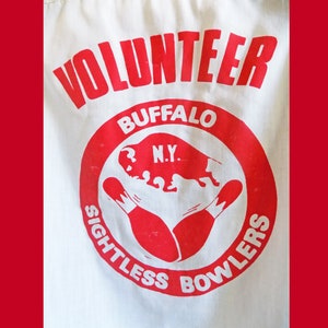 Op de afbeelding: Wit shirt met een rood cirkelvormig logo met de tekst "Volunteer, Buffalo N.Y., Sightless Bowlers". Het logo bevat ook een rood silhouet van een buffel en twee rode kegels.