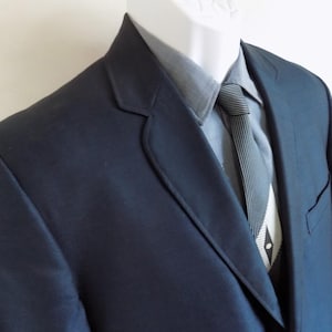 May include: A navy blue suit jacket with a matching tie and a light gray dress shirt. The tie has a black and white geometric pattern. The suit is displayed on a mannequin.