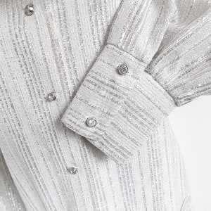 Puede incluir: Primer plano de una prenda de tejido plateado texturizado. La prenda presenta rayas verticales y está adornada con botones redondos y brillantes. Las mangas tienen puños abotonados con adornos similares. El diseño sugiere un estilo formal o elegante.