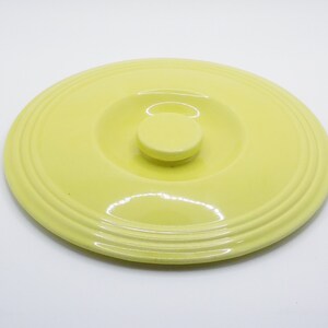 Puede incluir: Una tapa de cerámica amarilla con una perilla elevada en el centro. La tapa tiene un acabado liso y brillante y un borde ligeramente redondeado.