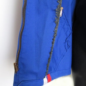 Puede incluir: Una chaqueta azul con cremallera y un borde deshilachado. La chaqueta tiene una franja blanca y roja en la parte inferior.