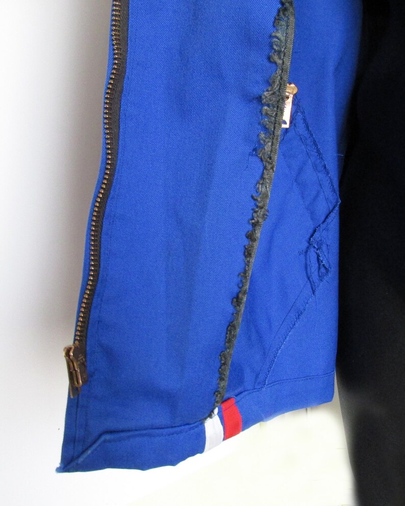 Puede incluir: Una chaqueta azul con cremallera y un borde deshilachado. La chaqueta tiene una franja blanca y roja en la parte inferior.